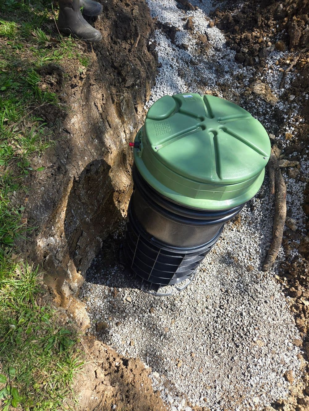 Green composting bin installed in a gravel-lined trench with surrounding soil and grass.