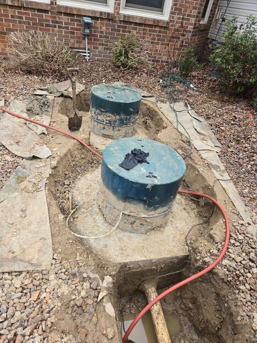 Excavated area with two blue septic tank covers and a shovel, surrounded by gravel and landscaping.