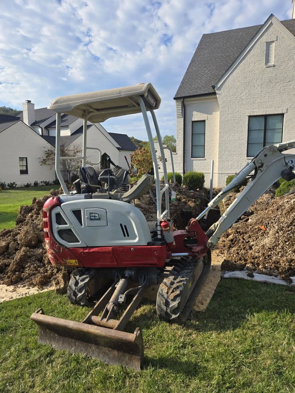 Mini excavator parked near a house, surrounded by dirt and construction materials.