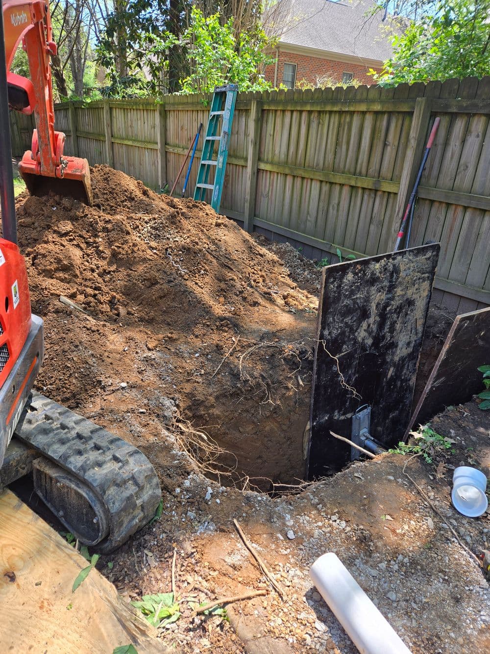 Excavation site with a large dirt mound, a digger, and a deep hole next to a wooden fence.