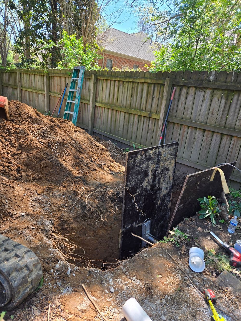 Excavation site with a deep hole, dirt mounds, and ladder near a wooden fence.