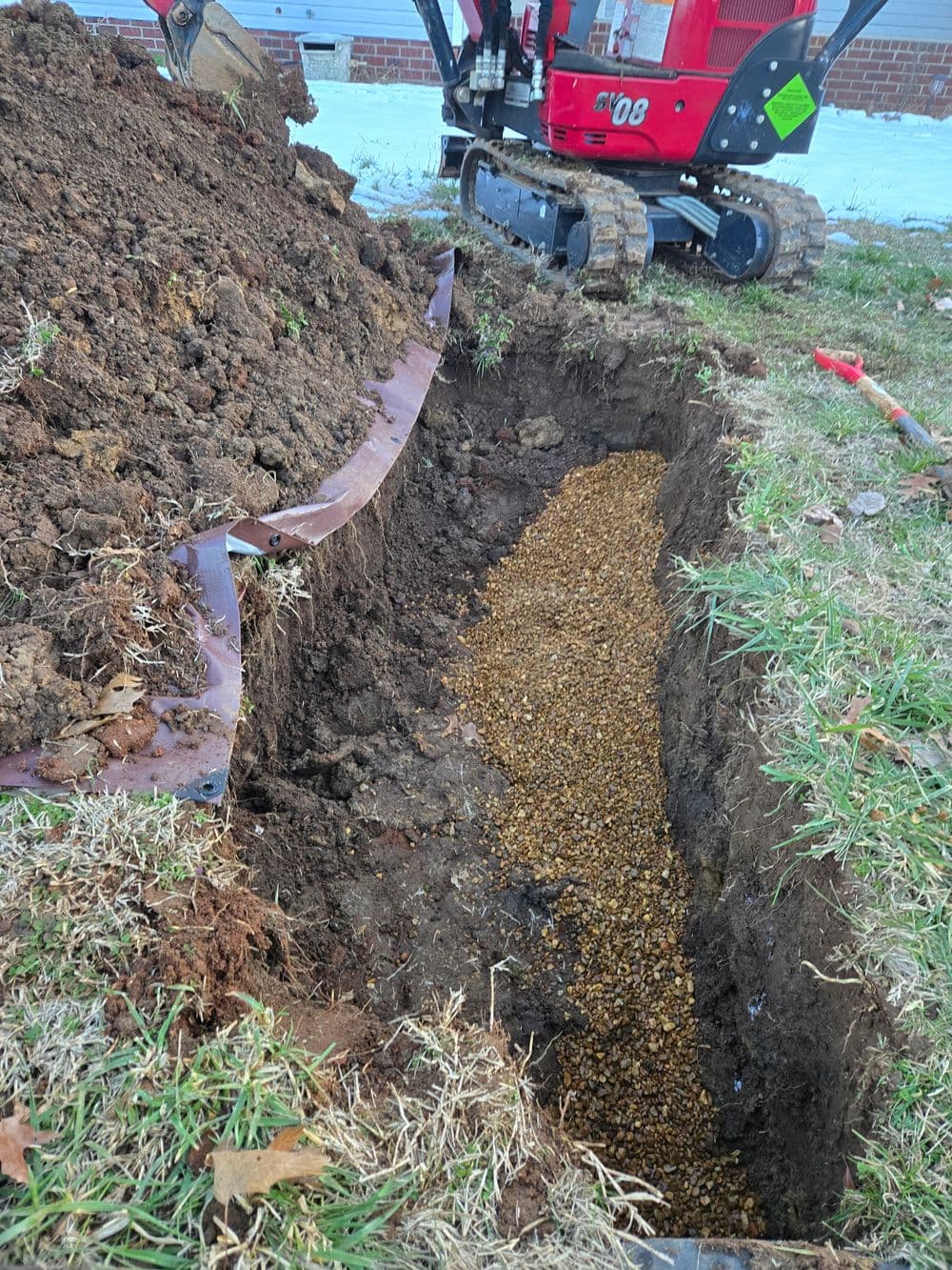 Excavated trench with gravel base and machinery in background for landscaping project.